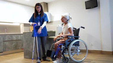 Hareketlilik bozukluğu olan yaşlı bir kadını destekleyen kendini adamış bir bakıcı, bir huzurevindeki fizik tedavi seansında tekerlekli sandalyeden kalkıp koltuk değnekleriyle yürümesine yardım ediyor.