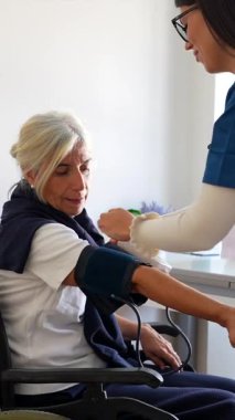 Klinikte yapılan rutin kontroller sırasında tekerlekli sandalyedeki yaşlı bir kadının tansiyonunu ölçmek için tonometre kullanan bir tıp uzmanı, kardiyovasküler sağlık denetimini...