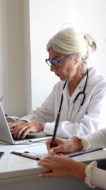 Genç bir hemşire meslektaşı modern bir klinikteki tıp eğitimi sırasında not alırken, steteskopla dizüstü bilgisayarda yazı yazan kıdemli bir kadın doktor.