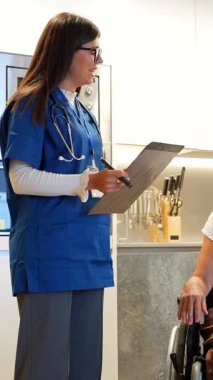 Mavi üniformalı profesyonel bir kadın doktor, mutfakta ev ziyareti sırasında tekerlekli sandalyede oturan mutlu ve şaşırmış bir yaşlı kadınla pozitif test sonuçlarını paylaşıyor.