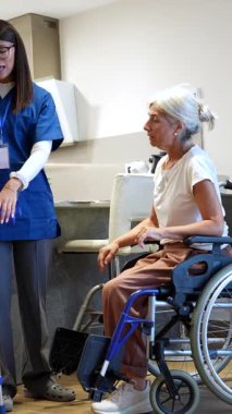 Son sınıf bir kadına iyileşmesi için yardım eden destekleyici bir bakıcı, bir huzurevindeki fizik tedavi seansında tekerlekli sandalyeden kalkmasına ve koltuk değnekleriyle yürümesine yardımcı oluyor.