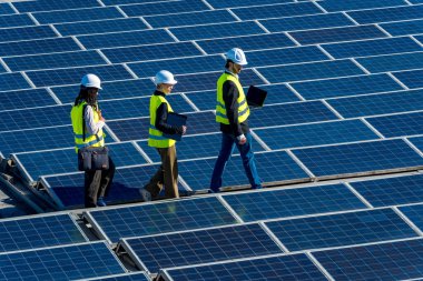 Sert şapka ve yelekli mühendisler büyük bir güneş panelinde yürüyorlar, kurulumu denetliyorlar ve temiz enerji üretim sisteminin verimliliğini sağlıyorlar.