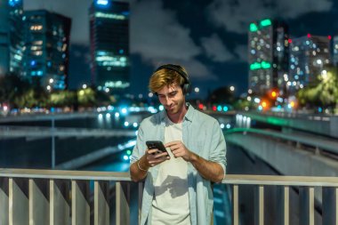 Kulaklık takan ve sıradan kıyafetler giyen genç bir adam, gece bir köprüde durmuş, akıllı telefonuyla kentsel bir arka planda gülümsüyor ve iletişim kuruyor.