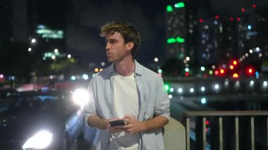 Yakışıklı genç adam endişeyle gece bir caddede birini bekliyor, cep telefonunu kontrol ederken etrafa bakıyor, arka planda bulanık trafik ışıkları ve gökdelenler var.
