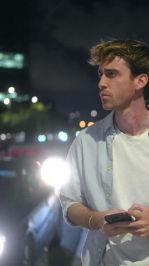 Ciddi bir beyaz adam gecenin bir yarısı şehir caddesinde birini bekliyor, elinde trafik ışıkları ve arka planda bokeh ile cep telefonunu tutarken ve kullanırken gergin bir şekilde etrafa bakıyor.