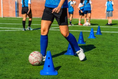 Kadın futbolcu yeşil bir sahada mavi antrenman konileri arasında top sürüyor. Takım arkadaşları arka planda antrenman yapıyor.