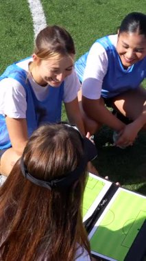 Profesyonel bayan futbol koçu yeşil alanda gülümseyen genç kadınlarla taktik ve stratejilerini tartışıyor, takım çalışmasını teşvik ediyor ve bir sonraki yarışma için hazırlık yapıyor.