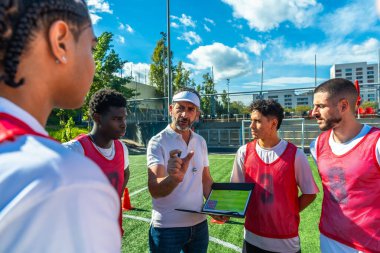 Futbol koçu, yeşil bir sahada çeşitli erkek oyunculara oyun stratejilerini açıklarken takım çalışmalarını, koçluklarını ve antrenman sırasında profesyonel planlarını gösteriyor.