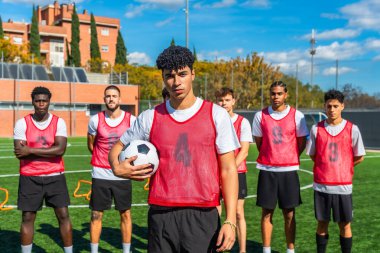 Kırmızı önlüklü çok ırklı gençler yeşil bir futbol sahasında durup antrenman veya maça hazırlanırken takım çalışması ve kararlılıklarını gösteriyorlar.