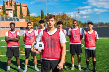 Koç, oyuncular ve sporcular futbol antrenmanına hazırlanıyor, güneşli bir açık hava oturumunda yeşil bir sahada antrenman yapıyor, takım çalışması ve kararlılığa odaklanıyor.