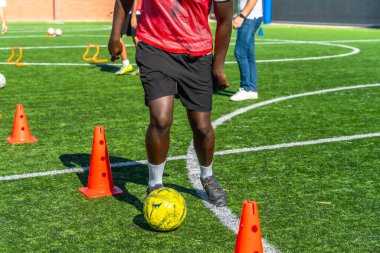 Genç oyuncu aktif olarak futbol talimi yapıyor ve pratik yapıyor. Yeşil yapay çim sahasında turuncu koniler etrafında top sürüyor.
