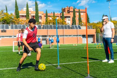 Antrenman oturumunda direkler arasında top sürmeye konsantre olan genç erkek sporcu bir koç gözlemliyor ve diğer oyuncular arka planda