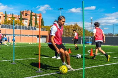 Kırmızı yelekli genç adam yeşil bir çim sahası üzerinde top sürme teknikleri öğreniyor, koni ve kutupların etrafında geziniyor diğer oyuncularla birlikte açık hava antrenmanı yapıyor.