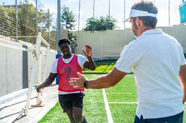 Genç erkek futbolcu, bir spor antrenmanı sırasında motivasyon, takım çalışması ve akıl hocalığını sembolize ederek sahada yetişkin koçuna beşlik çakıyor.