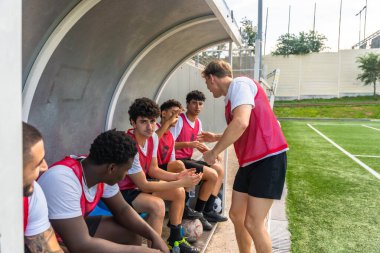 Gençlik futbol koçu, bir antrenman veya oyun sırasında spor sahasında çok ırklı erkek takım üyeleriyle strateji tartışıyor ve takım çalışmasını ve rehberliğini vurguluyor.