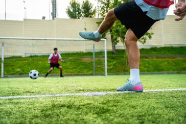 Futbolcunun bacağı, yeşil bir yapay çim sahasında penaltı atışında topa vuruyor. Kaleci arka planda bekliyor.