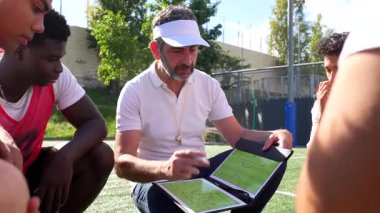 Bir futbol sahasında oturan erkek antrenör, çok ırklı genç oyunculardan oluşan takımına bir pano üzerinde oyun planını açıklarken dikkatle talimatları dinliyor.