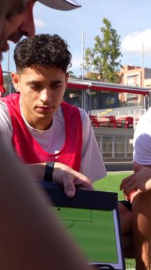 Erkek koç, güneşli bir günde futbol sahasında antrenman yaparken bir grup genç futbolcuya not panosundaki yeni oyun planını anlatıyor.