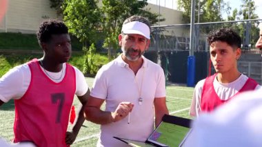 Orta yaşlı koç, maç planını açıklamak için bir tabletle birlikte bir tablet kullanıyor ve güneşli bir günde antrenman sırasında genç erkek futbolculardan oluşan bir takıma katılıyor.