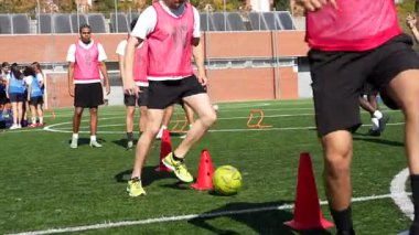 Pembe önlüklü genç erkek atletler futbol antrenmanı sırasında top sürme becerileri ve çeviklik pratiği yapıyor, sentetik bir sahada koçların gözetiminde kırmızı konilerde geziniyorlar.