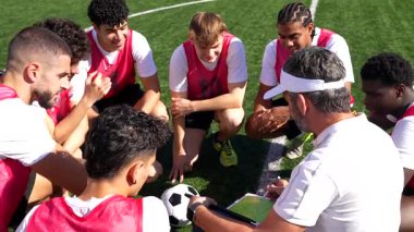 Erkek futbol koçu, çok ırklı genç oyunculardan oluşan takımına talimat veriyor. Bir futbol sahasında yeşil bir çember üzerinde otururken bir panoda oyun taktiklerini anlatıyor.