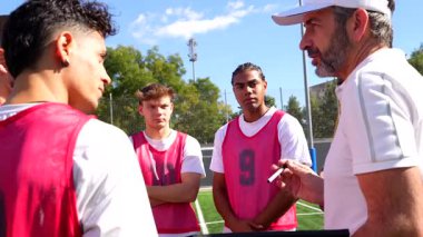 Profesyonel koç, güneşli bir günde antrenman sırasında çok kültürlü bir grup genç futbolcuya oyun stratejisi ve taktiklerini açıklarken takım çalışmasını ve akıl hocalığını teşvik ediyor.