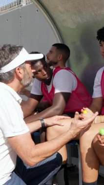 Olgun bir futbol menajeri, genç, çeşitli oyunculardan oluşan takımını, güneşli bir antrenman sırasında yedek kulübesinde oturarak strateji ve taktikleri açıklamaya teşvik ediyor.