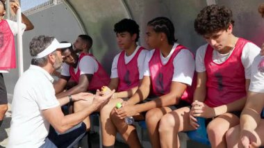 Orta yaşlı koç, güneşli bir antrenman sırasında yedek kulübesinde oturan oyunculara taktik ve motivasyon veriyor.