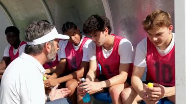 Profesyonel yetişkin koçu ara sırasında yedek kulübesinde oturuyor, konuşuyor ve genç oğlanlardan oluşan gençlik futbol takımına talimatlar veriyor, açık bir strateji ile onları maç için motive ediyor.