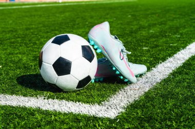Futbol topu ve futbol kramponları yeşil suni çimen sahasında oturuyorlar, güneşli koşullar altında canlı oyun sahasının beyaz sınır çizgisine yakın.