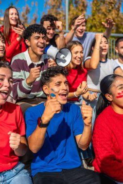 Genç çeşitli taraftarlar yoğun bir şekilde bir spor maçı izliyor, yüzlerini boyayıp kutlama yapıyorlar ve megafonla takım ruhunu ve stadyumdaki kolektif heyecanı gösteriyorlar.