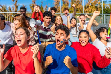 Çeşitli genç yetişkin ve arkadaşlardan oluşan enerjik bir grup takımlarına heyecan ve destek gösteriyor, yüksek sesle tezahürat yapıyor ve bir spor stadyumu veya arenasında zaferi kutluyorlar.