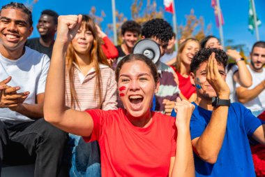 Çeşitli gençler bir stadyumda coşkuyla tezahürat yapıyorlar, takım renkleri giyiyorlar ve yüzlerini boyuyorlar ve bir spor müsabakasında en sevdikleri takıma hevesle destek veriyorlar.