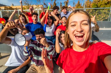 Genç taraftarlar, güneşli stadyum tribünlerinde tezahürat yapıyor ve selfie çekiyorlar. Heyecan verici bir maç sırasında takımlarını neşe, enerji ve dostlukla kutluyorlar.