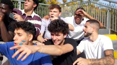 Stadyum tribünlerinde oturan, heyecanlanan, tezahürat yapan, bağıran ve güneşli bir günde tutkulu bir şekilde maç seyreden çok ırklı bir grup genç erkek arkadaş.