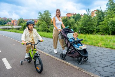 Genç bir anne küçük oğluyla bebek arabasını iterken, büyük oğlu kaskla bisiklet sürerken, güneşli bir günün tadını çıkarırken, parkta kahkaha ve beraberlikle dolu.
