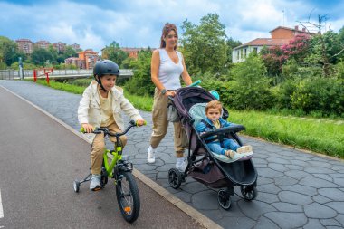 Anne, şehir parkında sakin bir gezinti yaparken, büyük oğlu bisiklete binip aile bağlarını ve aktif bir yaşam tarzını desteklerken, bebeğiyle bebek arabasını iterek eğleniyor.