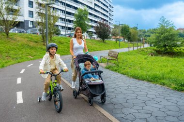 Anne, yeni yürümeye başlayan çocukla bebek arabasını itiyor ve şehir parkında bisiklet sürüyor. Ev binalarının yanında, dışarıda ailecek vakit geçiriyor.