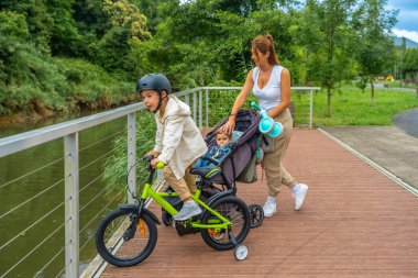 Parktaki bir nehrin kenarında bisiklet süren diğer oğlu güneşli bir günün tadını çıkarırken genç bir anne, içinde bebekle bebek arabasını itiyor.
