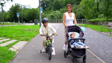 Mutlu anne, iki çocuğuyla parkta yürüyor. Biri bisiklet sürüyor. Bisiklet tekerlekleri, miğferi ve diğeri bebek arabasıyla güneşli bir günün tadını çıkarıyor.