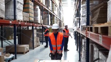 Büyük bir dağıtım merkezinin envanterini yöneten lojistik işçilerden oluşan çeşitli bir ekip, bir çalışanın bir palet krikosuyla kutuları taşırken, diğerleri yüksek metal raflardaki stokları kontrol ediyor.