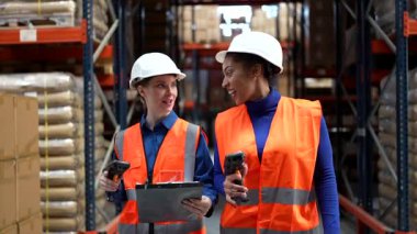 Lojistik deposunda barkod tarayıcıları ve envanteri yönetmek ve sevkiyat detaylarını tartışmak için bir pano kullanan çok ırklı kadın meslektaşlar.