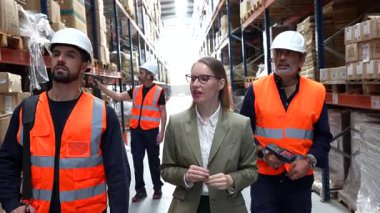 Kadın yönetici depo koridorlarında yürüyor, takım üyeleriyle envanter ve lojistiği tartışıyor, rafları işaret ediyor ve stokları koordine ediyor, teftiş sırasında görünen güvenlik kaskları