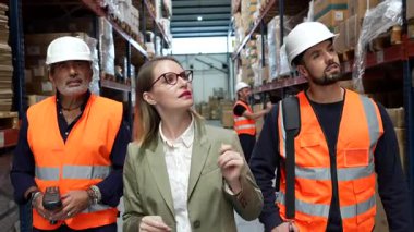 Profesyonel kadın yönetici, iki erkek çalışanla birlikte büyük bir depoda envanterleri kontrol ediyor ve kutularla dolu uzun raflar arasında ürün dağılımını tartışıyor.