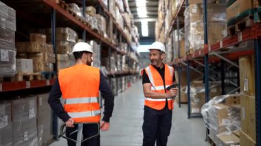 Sıkı bir şapka ve güvenlik yeleği giyen odaklanmış bir erkek depo çalışanı paketlerdeki barkodları tararken, arka planda palet kriko kullanan bir meslektaşı da verimli bir envanter kontrolü sağlıyordu.