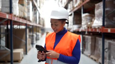 Dağıtım merkezindeki stokları, nakliyatları ve stokları kontrol etmek için yığılmış kutuların üzerindeki barkodları tarayan çelik yelek ve çelik yelek giyen kadın lojistik işçisi.