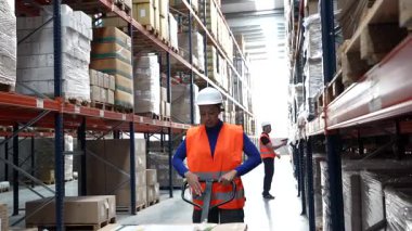 Kadın lojistik çalışanı, arka plandaki envanteri kontrol ederken karton kutularla dolu bir manüel palet çıkarıyor. Büyük bir dağıtım deposunda birlikte çalışıyoruz.