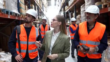Kadın depo müdürü, erkek işçilerle dolu büyük bir depo koridorunda yürüyor, raflardaki envanterleri inceliyor ve nakliye ve dağıtım için lojistik konularını tartışıyor.
