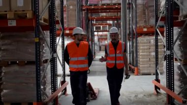 Güvenlik yelekleri ve başlıklı iki erkek arkadaş büyük bir sanayi deposu koridorunda yürüyorlar, uzun raflar arasında çalışma süreçlerini, lojistik yönetimini ve envanter kontrolünü tartışıyorlar.