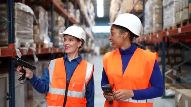 Güvenlik yelekleri ve başlık takan iki farklı kadın depo çalışanı raflar arasında yürüyor, el bilgisayarlı bir aletle barkodları eşleştiriyor ve tarıyorlar.
